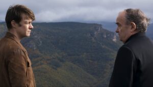 A young man and a priest talk to each other while standing on a rural cliff overlooking a green, hilly landscape.