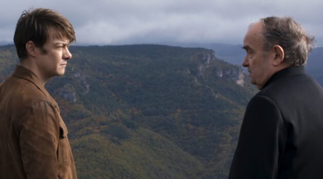 A young man and a priest talk to each other while standing on a rural cliff overlooking a green, hilly landscape.