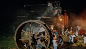 Peasant villagers surround a gigantic threshing wheel as a fire burns in the background.