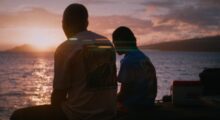 Two people sit on a beach at sunset.