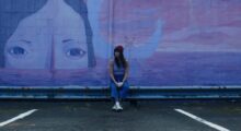 A woman in a red beanie leans against a wall with a mural of a woman's enormous head poking above ocean waves.