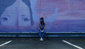 A woman in a red beanie leans against a wall with a mural of a woman's enormous head poking above ocean waves.