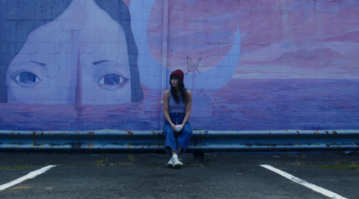 A woman in a red beanie leans against a wall with a mural of a woman's enormous head poking above ocean waves.