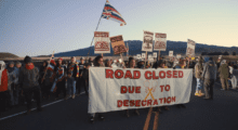 A group of Hawaiian protesters holds a protest on a road while holding a banner and flags.