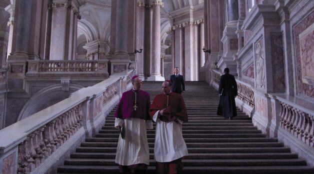 Two Catholic cardinals walk down a stairway in an elaborate building.