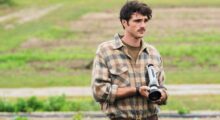 A man with a mustache holds a movie camera in an open field.