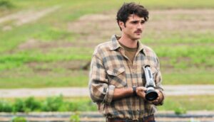 A man with a mustache holds a movie camera in an open field.