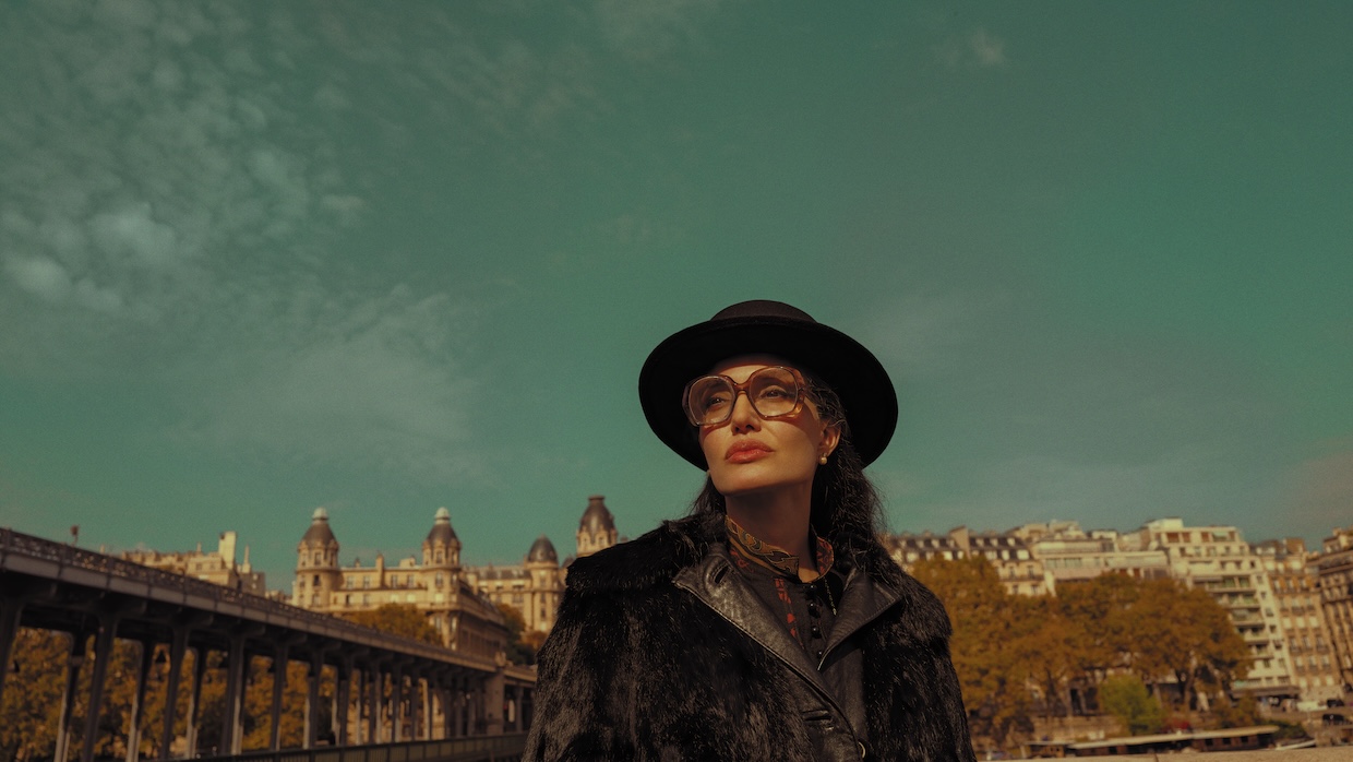 A woman wears a hat, glasses and coat while standing outside.