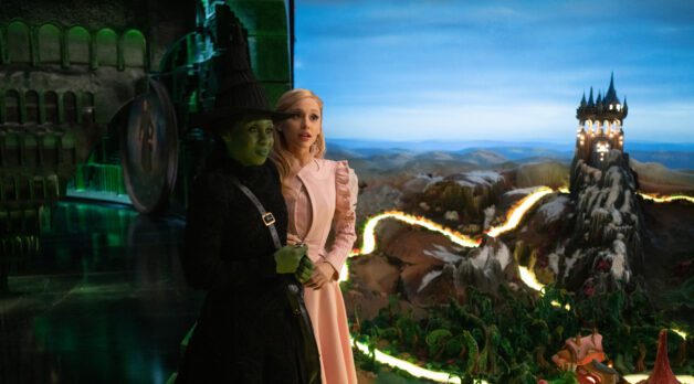 A green witch and white woman stand together against a very fake-looking background.