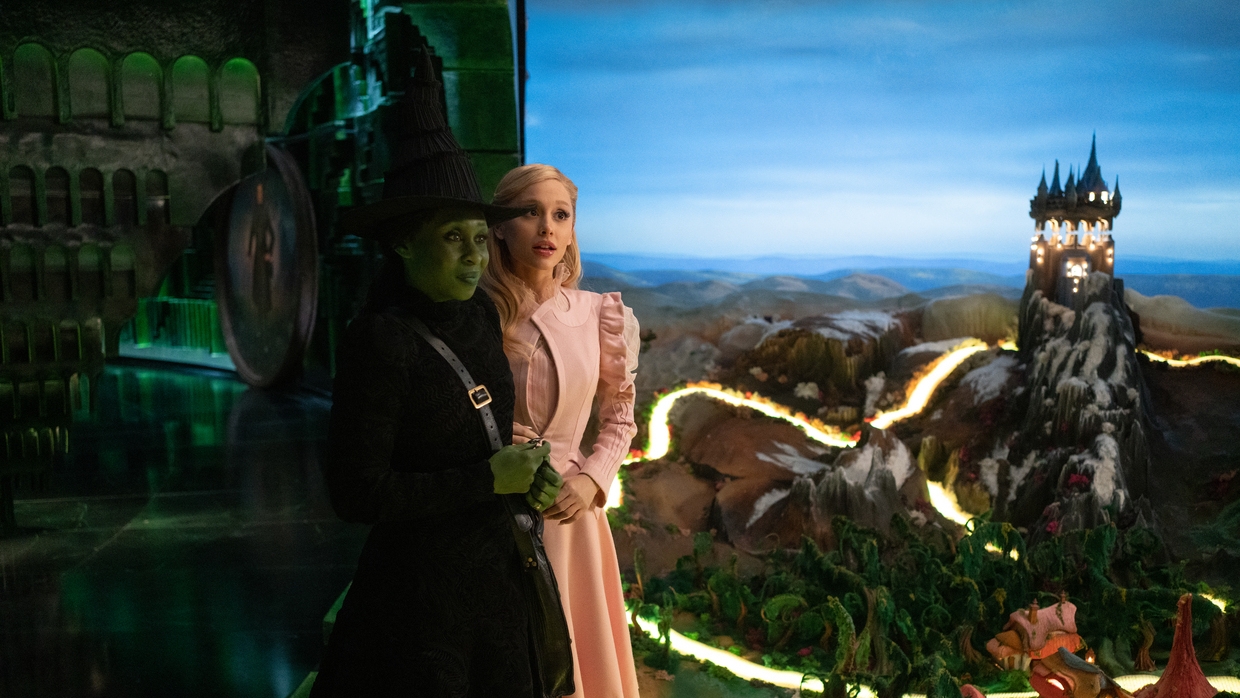 A green witch and white woman stand together against a very fake-looking background.