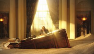 A man sits at a tilted office desk partially buried in sand.