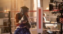 A woman in boxing gloves faces a camera on a film set.