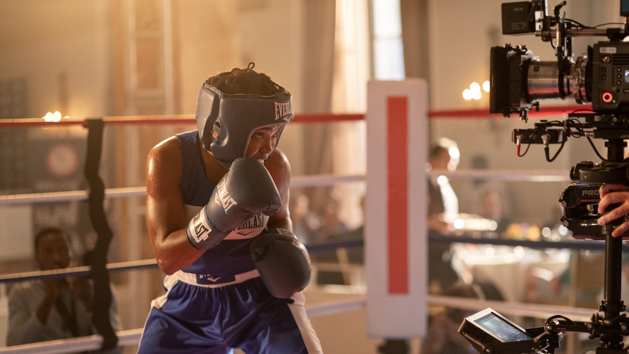 A woman in boxing gloves faces a camera on a film set.