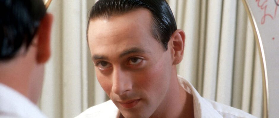 Paul Reubens looks into the mirror. He wears a white dress shirt and has his black hair slicked back.