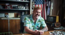 A white man in a tropical button-down is sitting at a desk.