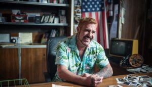 A white man in a tropical button-down is sitting at a desk.