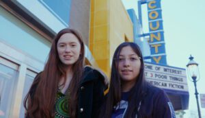 Two-shot of teenage girls on the sidewalk with a movie theater marquee in the background.