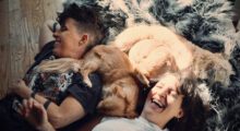 Two people are lying on the floor laughing while a small dog sleeps between them.