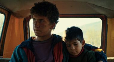 Two boys, one a teenager and one younger, are sitting inside. The younger is asleep.