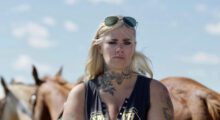 A blonde woman with heavy black eyeliner and a black tank top stands amongst brown horses.