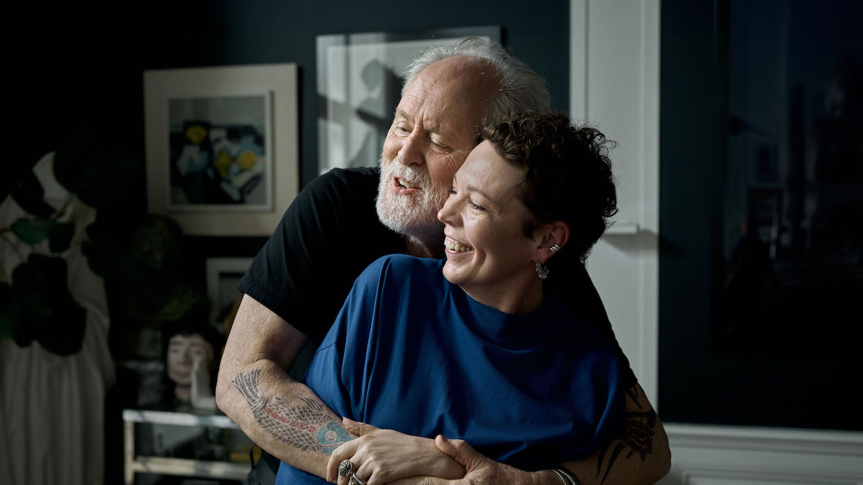 An older man with white hair and beard hugs a middle-aged woman with short black hair and a blue sweater.