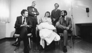 Black-and-white image of a woman in white on a hospital bed with four men in suits around her.