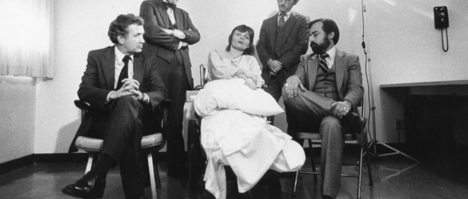 Black-and-white image of a woman in white on a hospital bed with four men in suits around her.