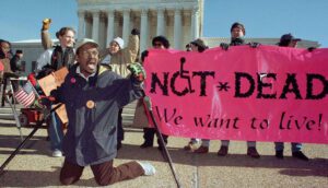 A disabilities rights protest with a banner reading "Not Dead, We Want To Live" visible.