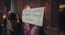 A masked person is holding a banner that reads, "Assisted Suicide: The World's Cheapest Healthcare"