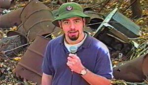 A young man wears a purple t-shirt, green bucket hat and sports a goatee. He holds a microphone and stands in front of barrels of indiscriminate waste.