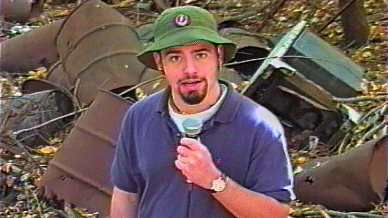 A young man wears a purple t-shirt, green bucket hat and sports a goatee. He holds a microphone and stands in front of barrels of indiscriminate waste.