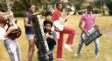 Several Black musicians are standing in the grass playing guitars and keyboards.