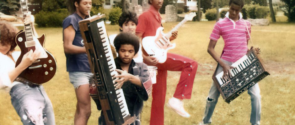 Several Black musicians are standing in the grass playing guitars and keyboards.