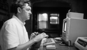 A white man is sitting at an old, CRT-monitor computer.