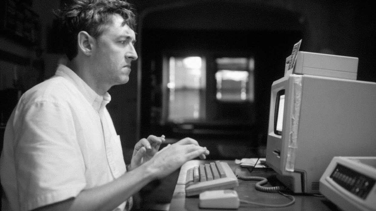 A white man is sitting at an old, CRT-monitor computer.