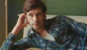 A white man in his 40s with dark hair and a blue plaid shirt leaning on a couch with cigarette