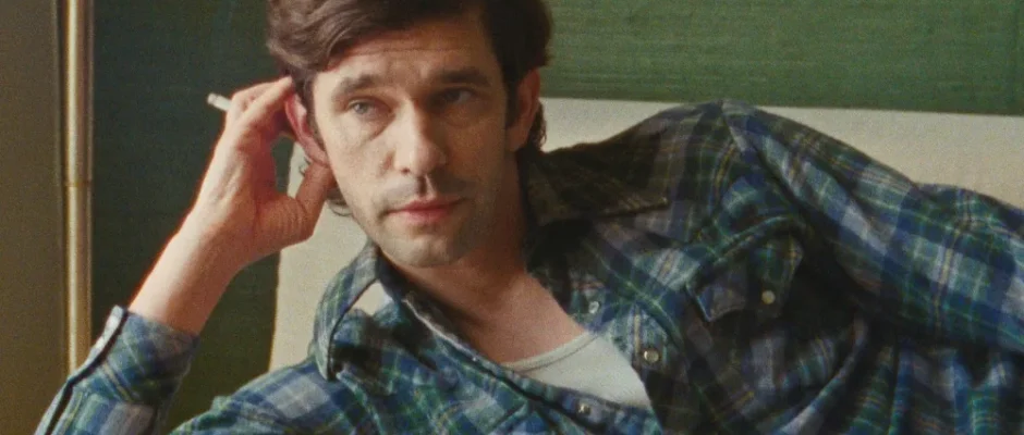 A white man in his 40s with dark hair and a blue plaid shirt leaning on a couch with cigarette