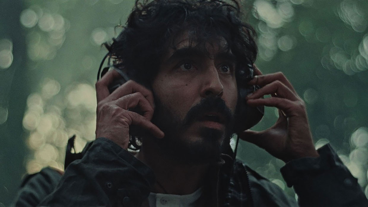 A man with black curly hair and a mustache listens to headphones with a concerned expression on his face.