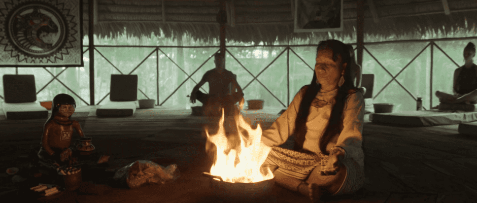 A woman sits near. small fire inside of a hut.