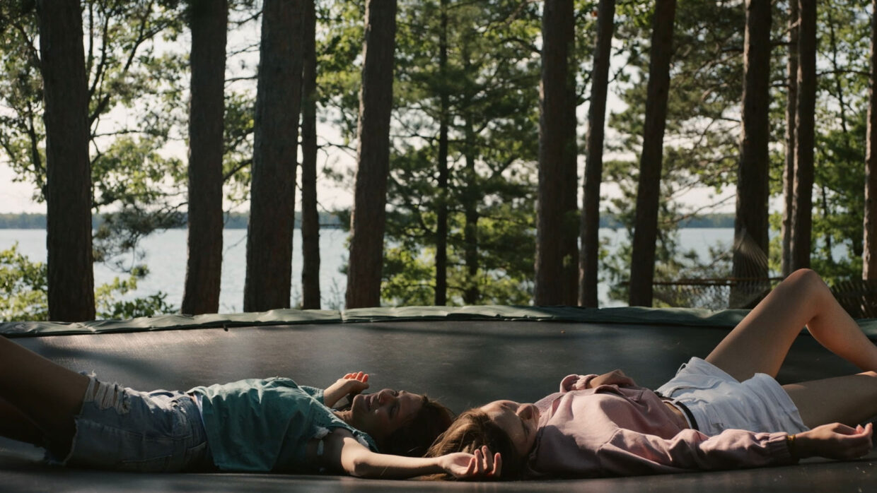 Two young people, one man and one woman, are lying on a trampoline. The ocean is visible in the background behind some trees.