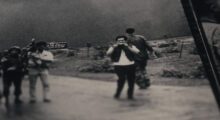 A black and white still of a camera crew and a military soldier passing behind them.