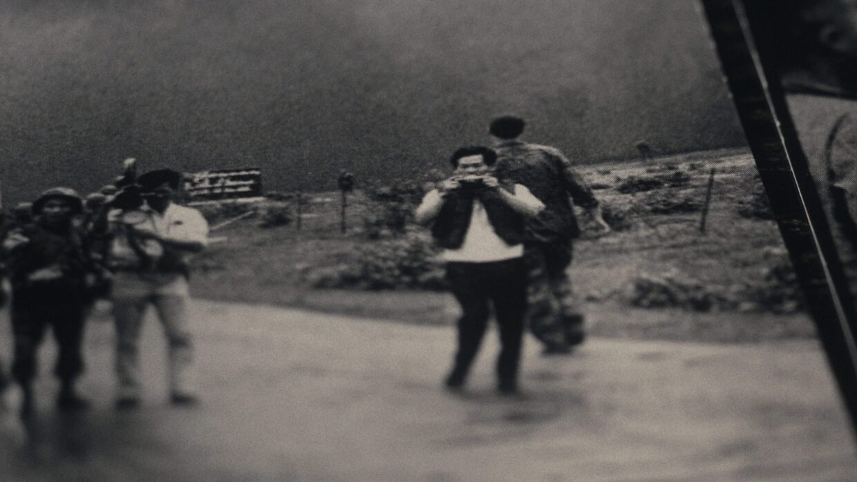 A black and white still of a camera crew and a military soldier passing behind them.