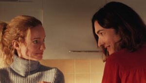 A blond woman in a grey turtleneck and a brunette in a red sweater look at each other.