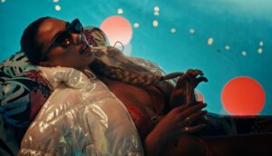 A woman wearing a white puffer jacket, red bikini top and sunglasses reclines amid a neon blue background.