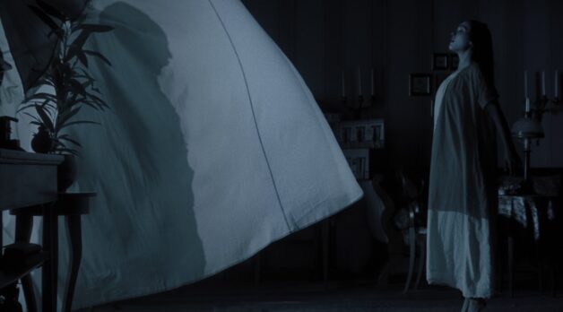 A man's caped silhouette is seen through a white window curtain with a woman standing next to it.