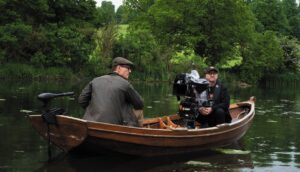 An actor in costume sits in a boat opposite a cinematographer with a camera.