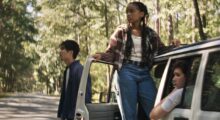 Three young people stand in the daylight woods next to their white car.