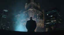 A man stands on a roof in the big city and stares into the night as steam rises before him.