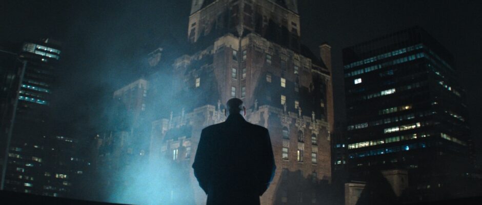 A man stands on a roof in the big city and stares into the night as steam rises before him.