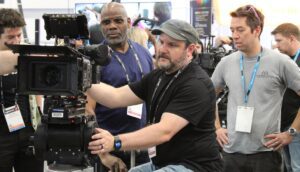 A group of man crowd around a camera on a trade show floor.